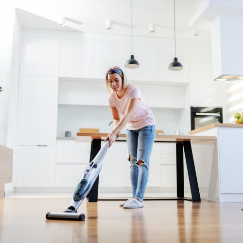 Full length of worthy caucasian blonde housewife using steamer to clean floor in living room.